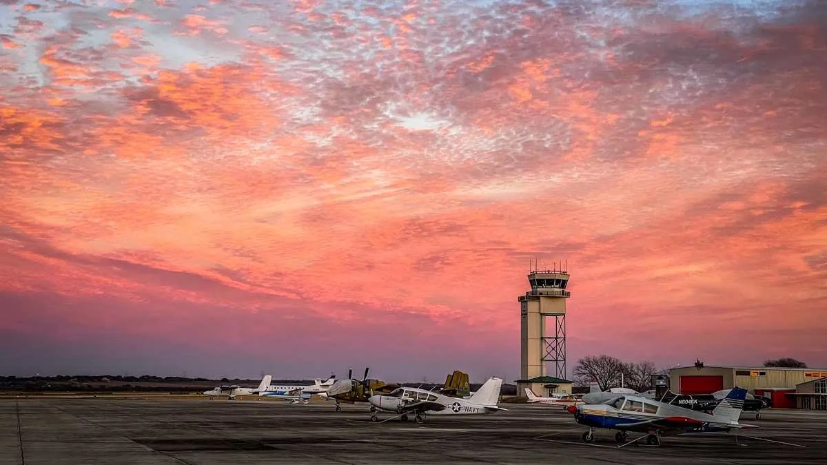 Aircraft parked beside the San Marcos Regional Airport control tower beneath a pink and orange sky